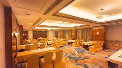 A corner view of the spacious banquet hall at Pride Plaza Ahmedabad, featuring multiple round tables with white tablecloths, set for a large gathering or event.